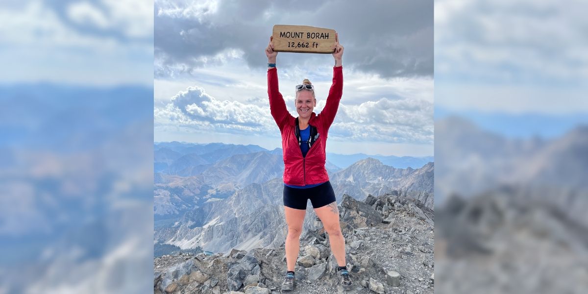 Woman in sportswear stands on top of mountain holding sign that reads &quot;MOUNT BORAH 12,662 ft&quot; triumphantly above her head