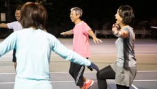 Group of people on a tennis court smile and laugh as they balance on one leg