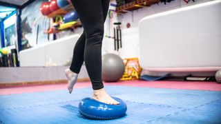 A barefoot woman balances on one leg on a balancing disc in a fitness studio